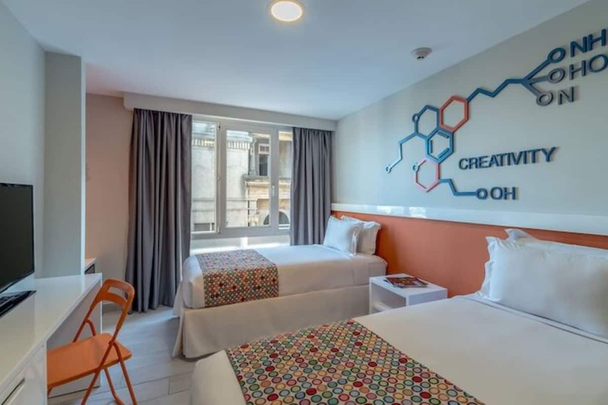 Ein Hotelzimmer mit zwei Betten, einem Schreibtisch mit Fernseher, einem Sessel und einem Fenster mit Vorhängen.