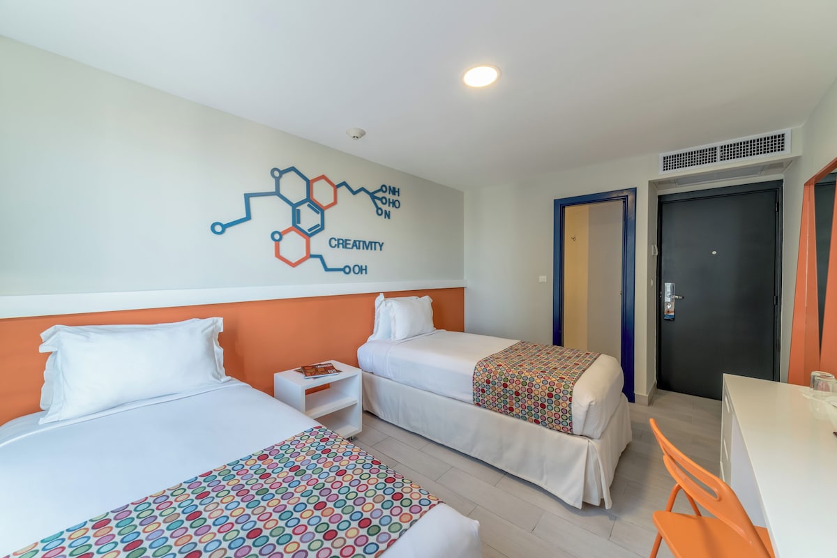 A hotel room with two beds, a desk, and a chair. There is a wall decoration with a molecular structure and the word "CREATIVITY".