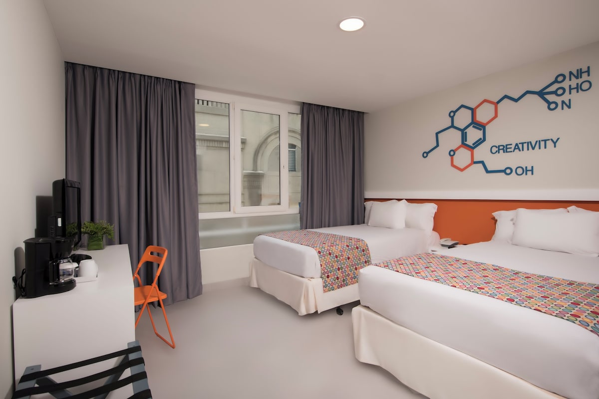 A hotel room with two beds, a desk with a coffee maker, a chair, and a window with curtains.
