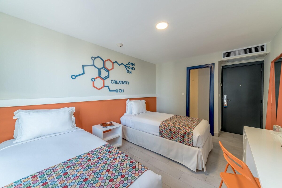 A hotel room with two beds, a desk, and a chair. There is a wall decoration with a molecular structure and the word "CREATIVITY".