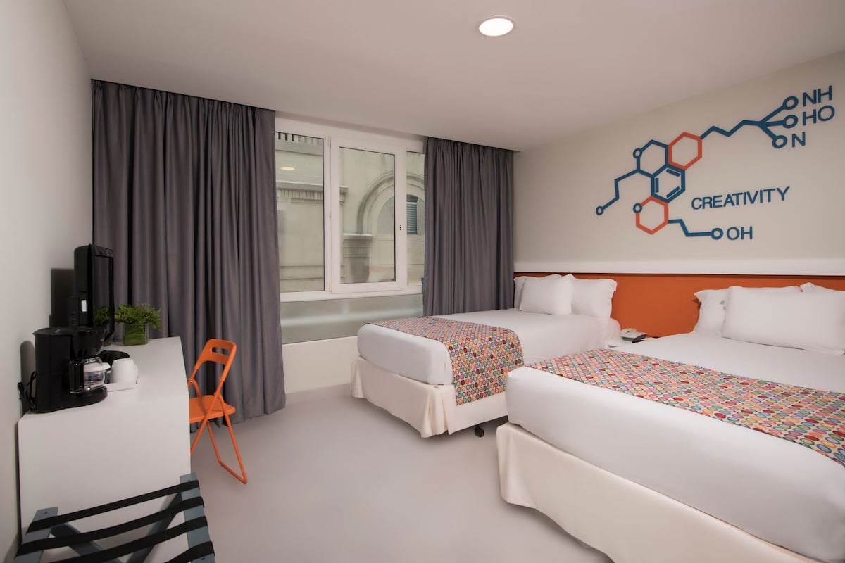 Habitación de hotel con dos camas, un escritorio con cafetera, una silla y un diseño temático en la pared.