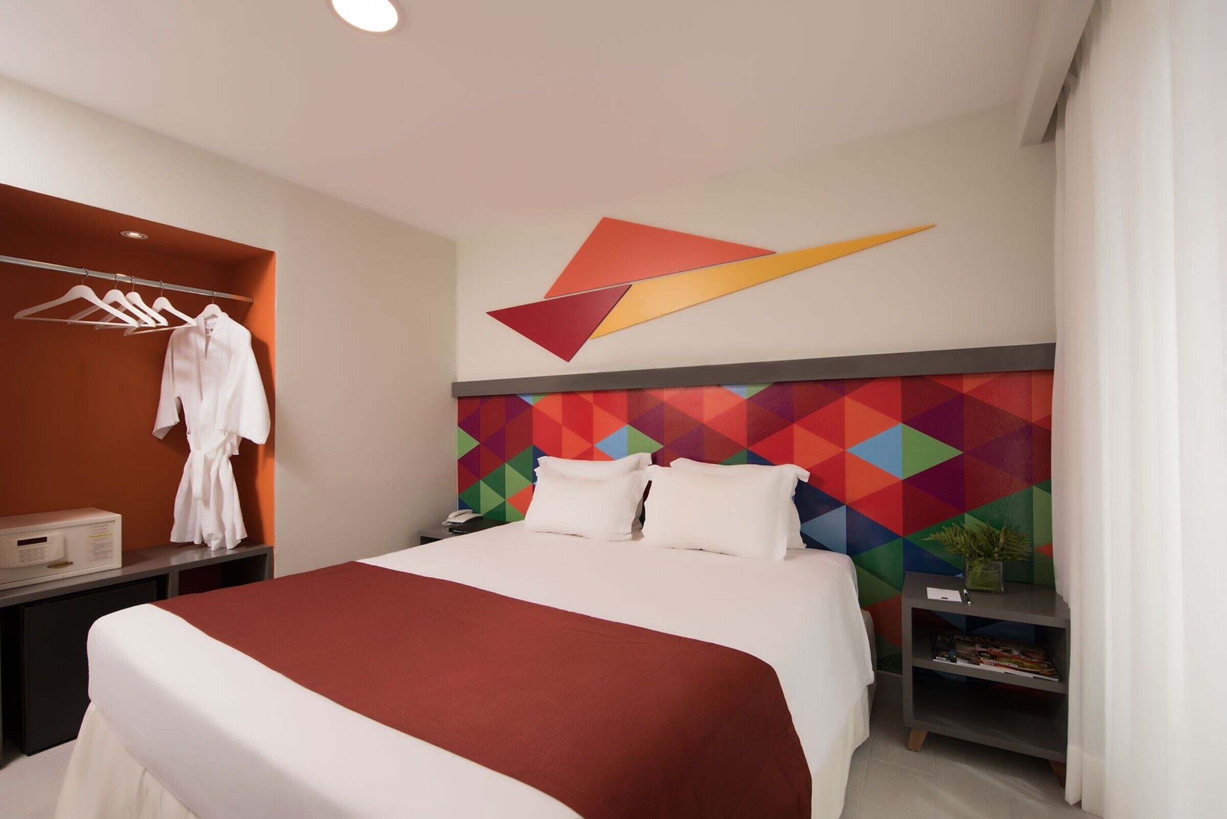A hotel room with a bed, a robe hanging, a nightstand, a wall-mounted geometric decoration, and a small plant on a side table.
