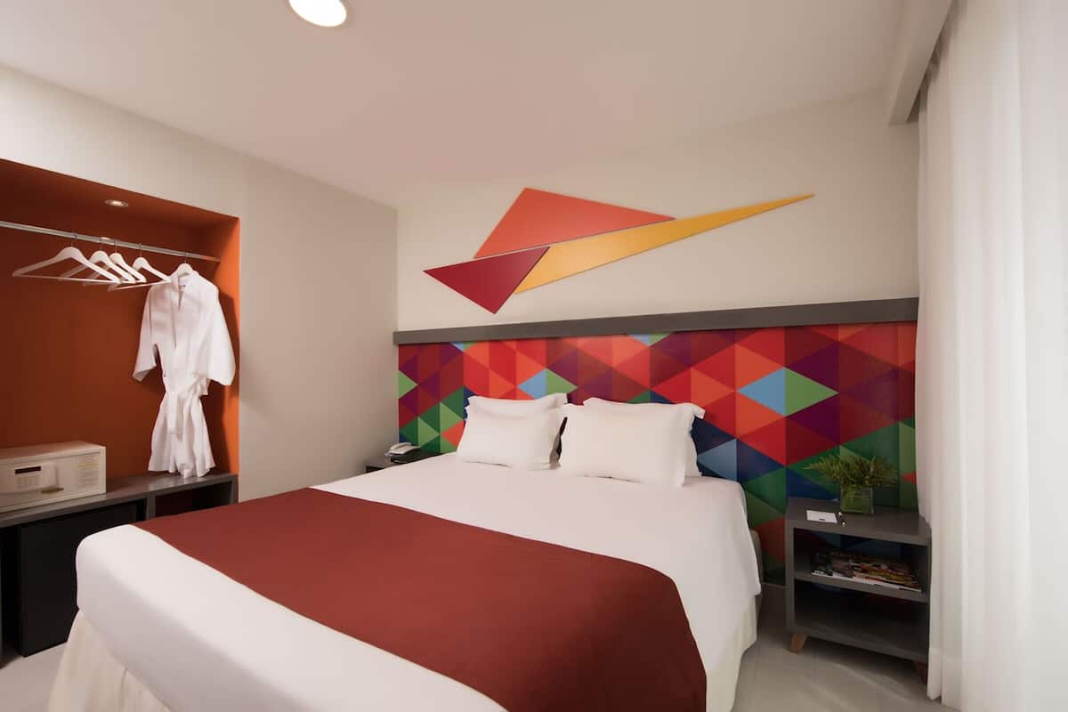 Ein modernes Hotelzimmer mit einem großen Bett, einem Bademantel an einem Gestell, einem Nachttisch mit Lampe und einem farbenfrohen, geometrischen Wanddesign.