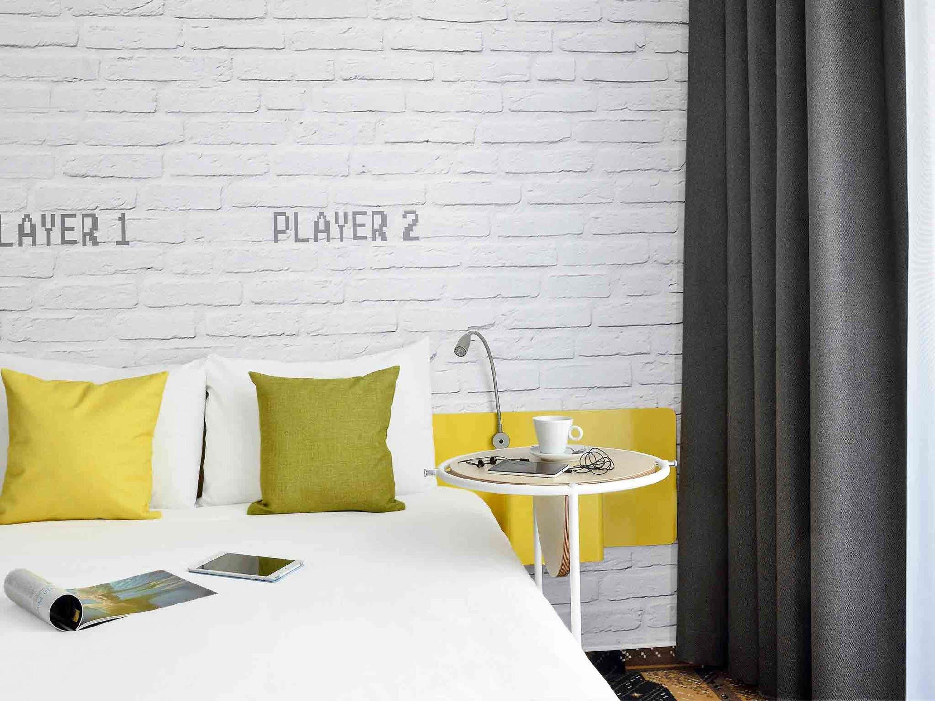 A hotel room with a white brick wall, two pillows, a bed, a small round table with a cup and a book, and a smartphone on the bed.