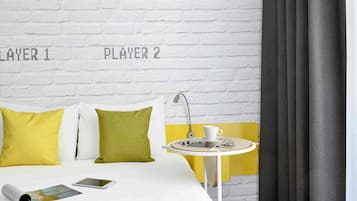 A hotel room with a white brick wall, two pillows, a bed, a small round table with a cup and a book, and a smartphone on the bed.
