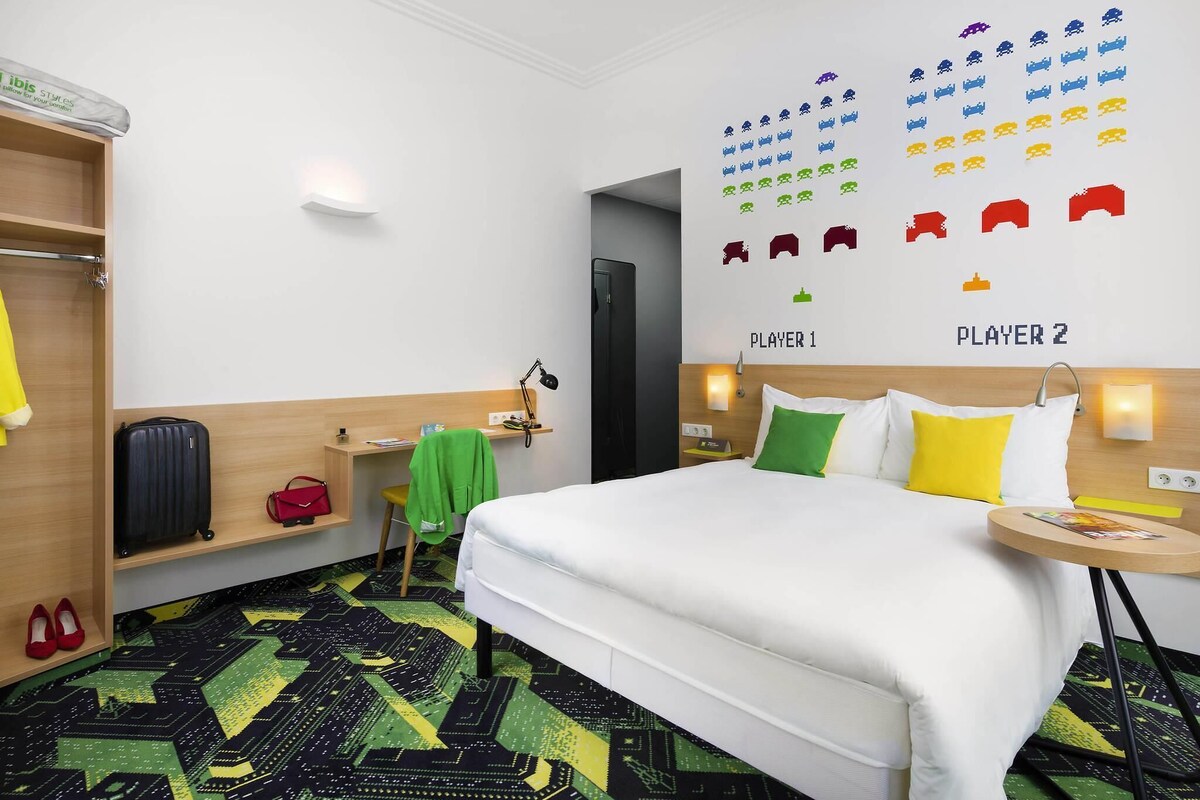 A hotel room with a bed, a desk, a chair, a wardrobe, and a wall with game icons.
