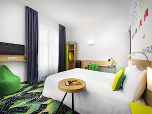 Hotelkamer met een bed, een klein tafeltje, een groene relaxfauteuil, een kledingkast, een televisie en een wand met kleurrijke decoraties.