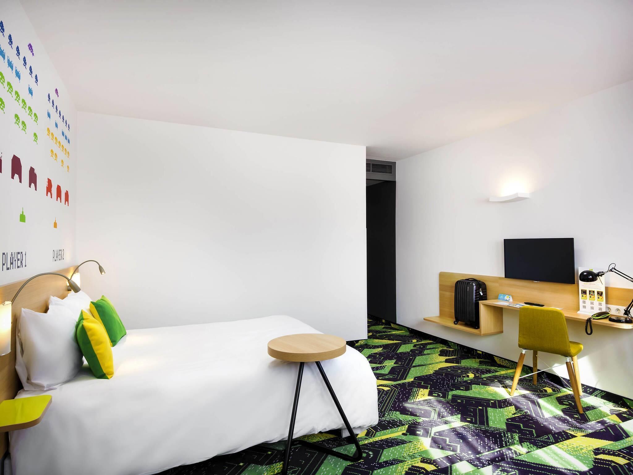 Hotellihuone, jossa on sänky, työpöytä, tuoli, televisio ja seinä, jossa on värikkäitä kuvakkeita.
