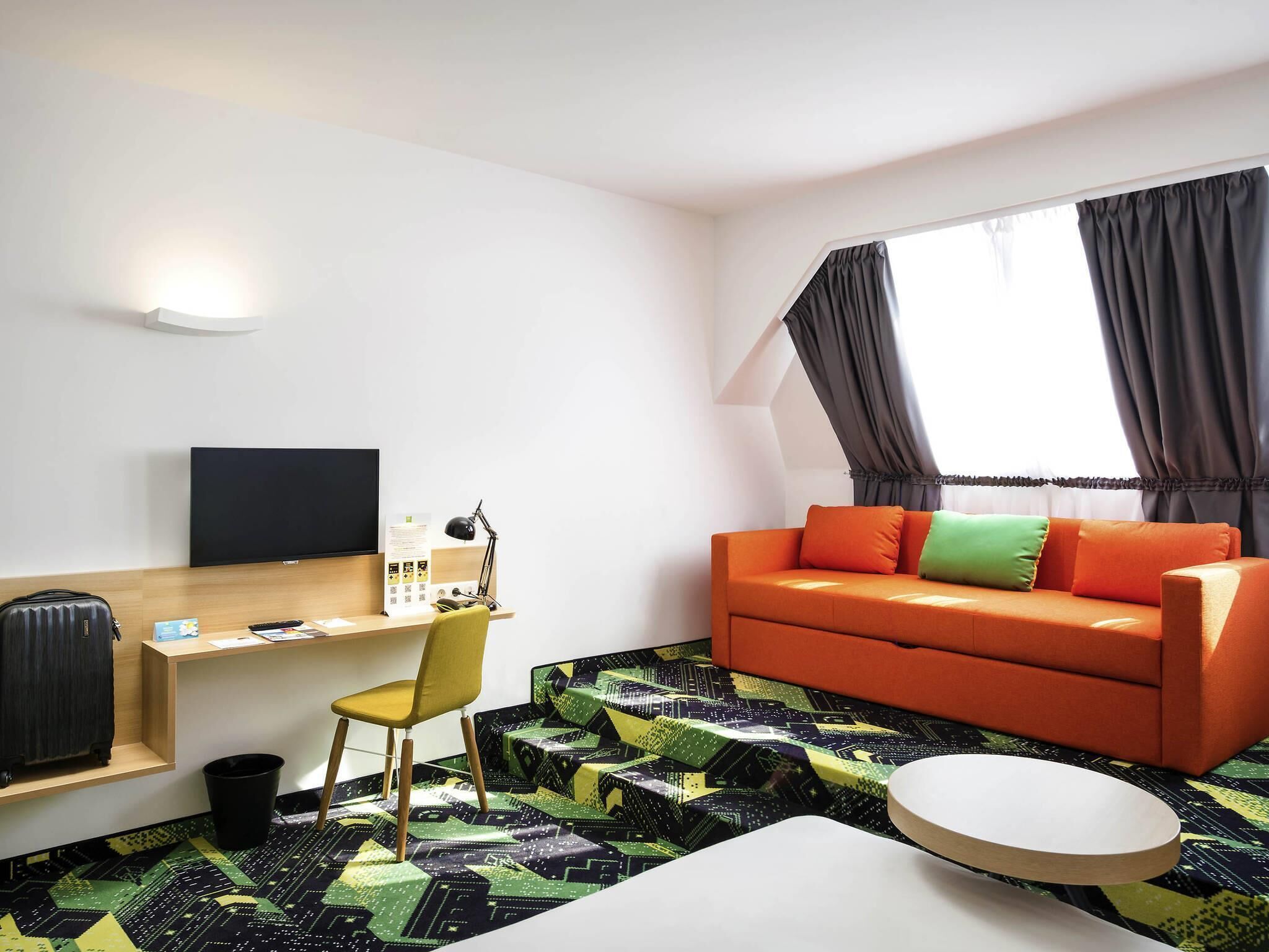 A modern hotel room with a wooden desk, a flat-screen TV, a sofa with cushions, a patterned rug, and a round coffee table.