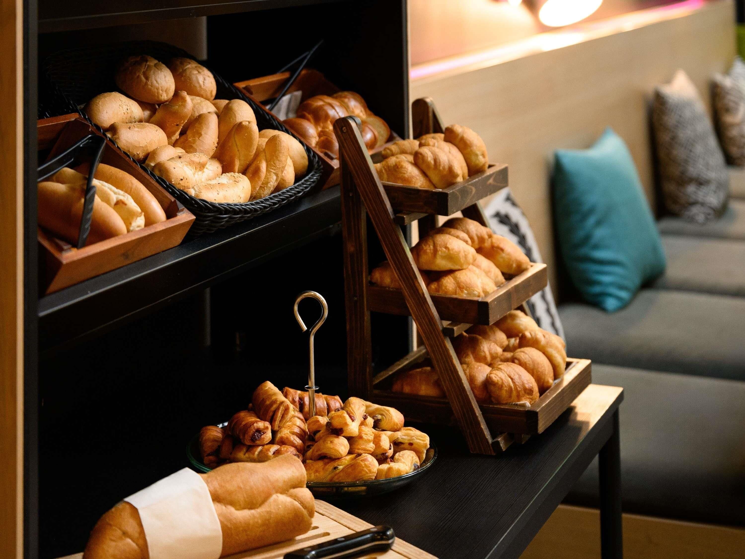 Uma mesa de buffet com diversos pães e doces, em ambiente moderno, com um sofá e almofadas ao fundo.