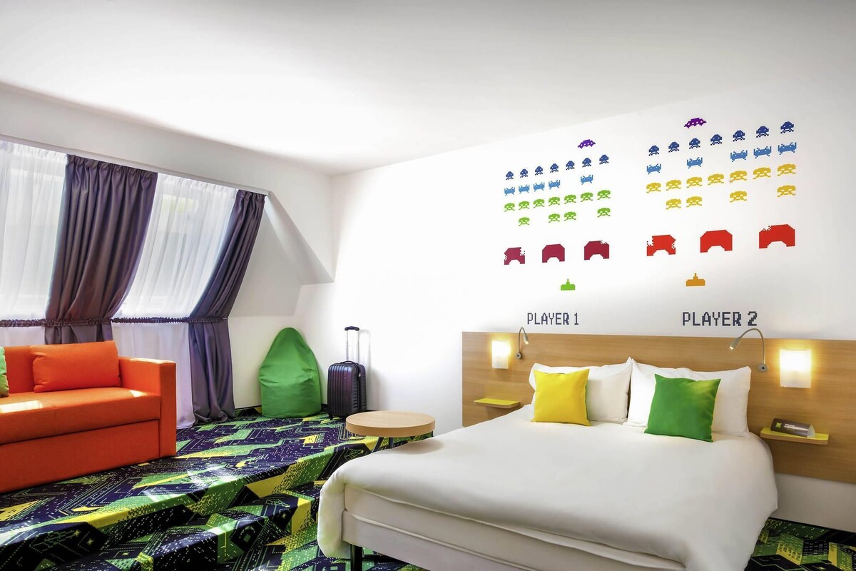A hotel room with a bed, a sofa, a green bean bag, a suitcase, and a wall with game icons.