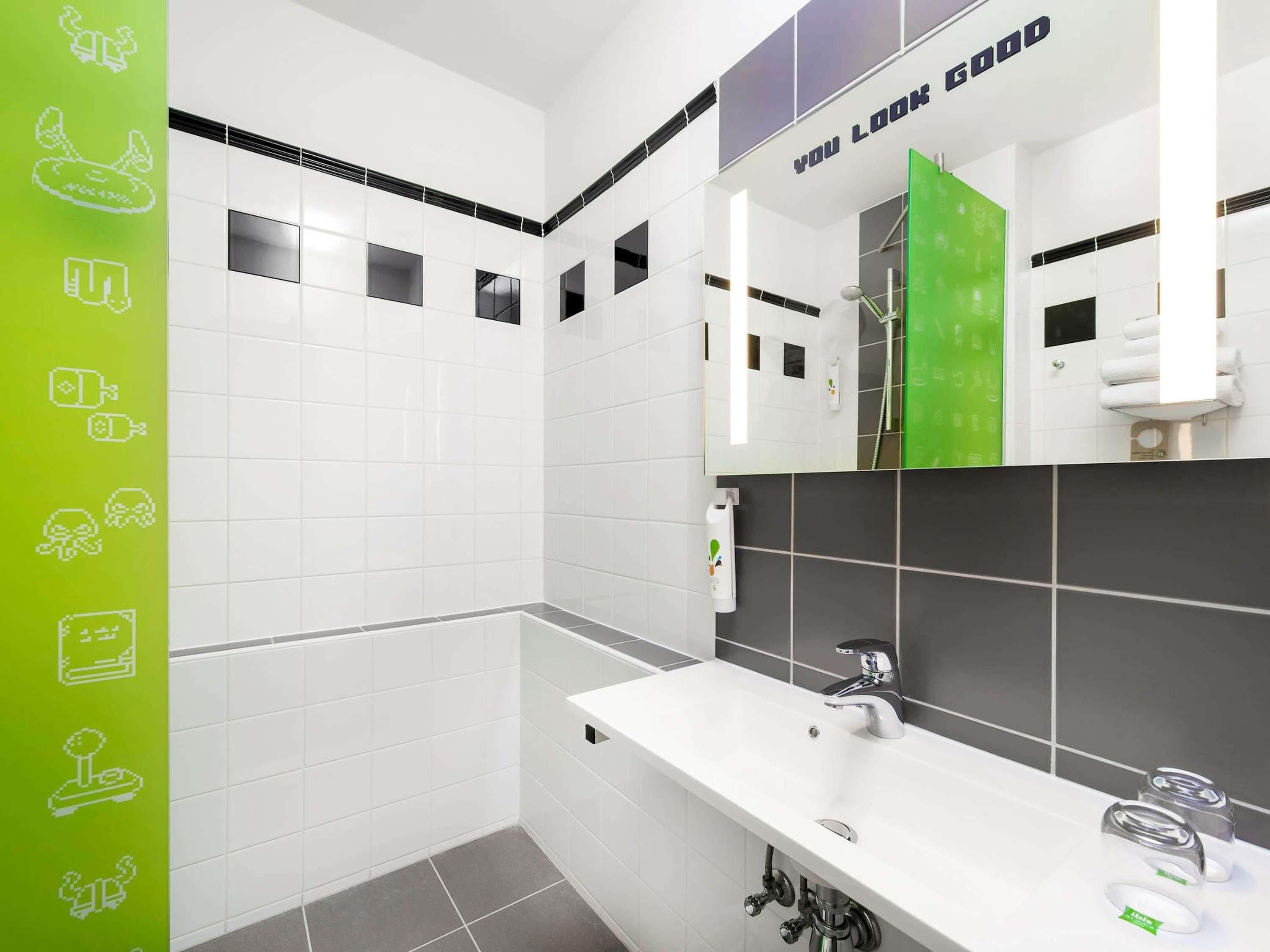 A modern bathroom with white tiles, a large mirror, a white sink, and a shower area.