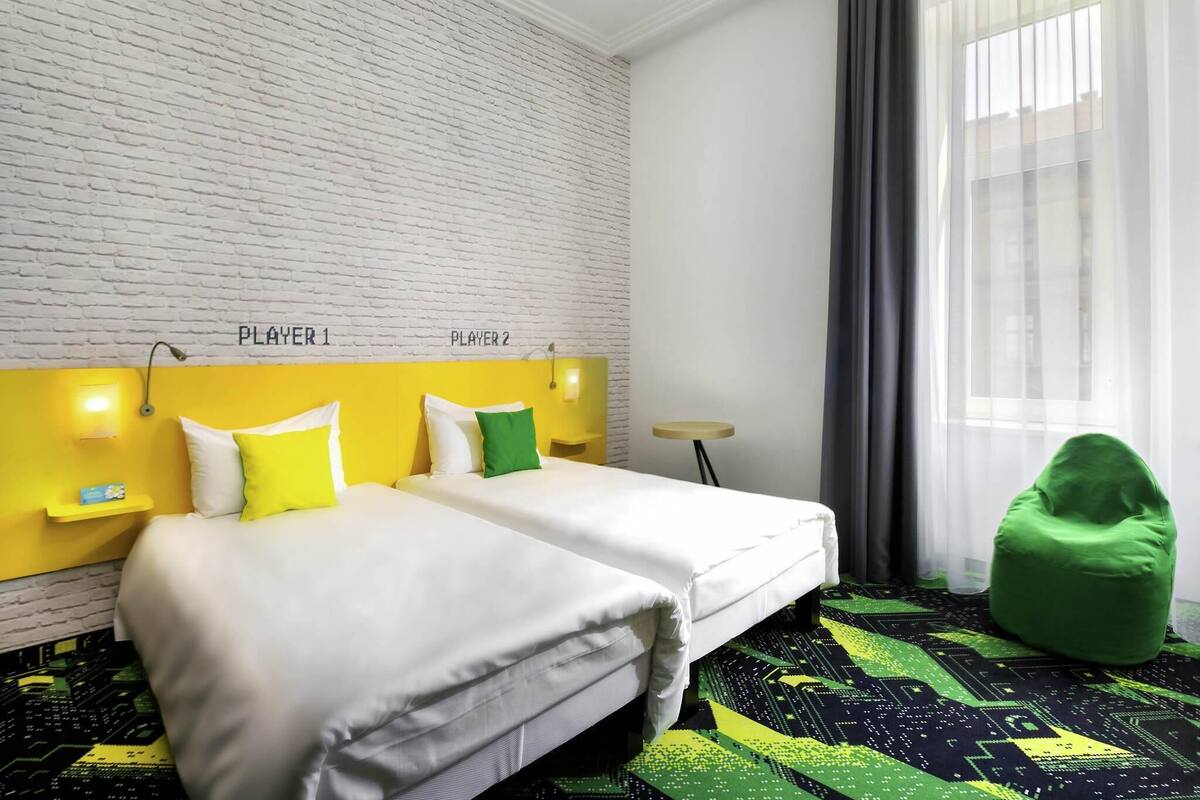 A hotel room with a white brick wall, two beds labeled 'PLAYER 1' and 'PLAYER 2', a green bean bag chair, and a patterned carpet.