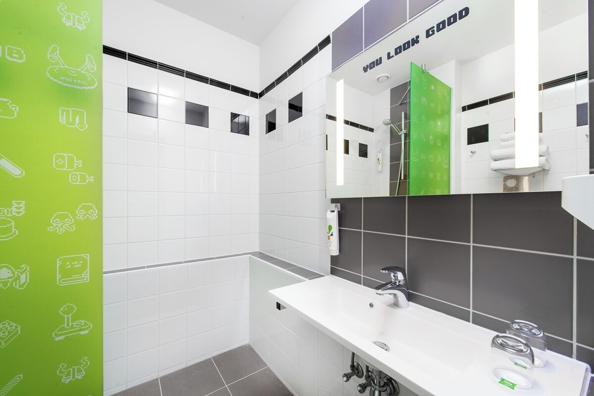 Un baño moderno con azulejos blancos y grises, un espejo grande, una ducha y un lavabo blanco con grifo.