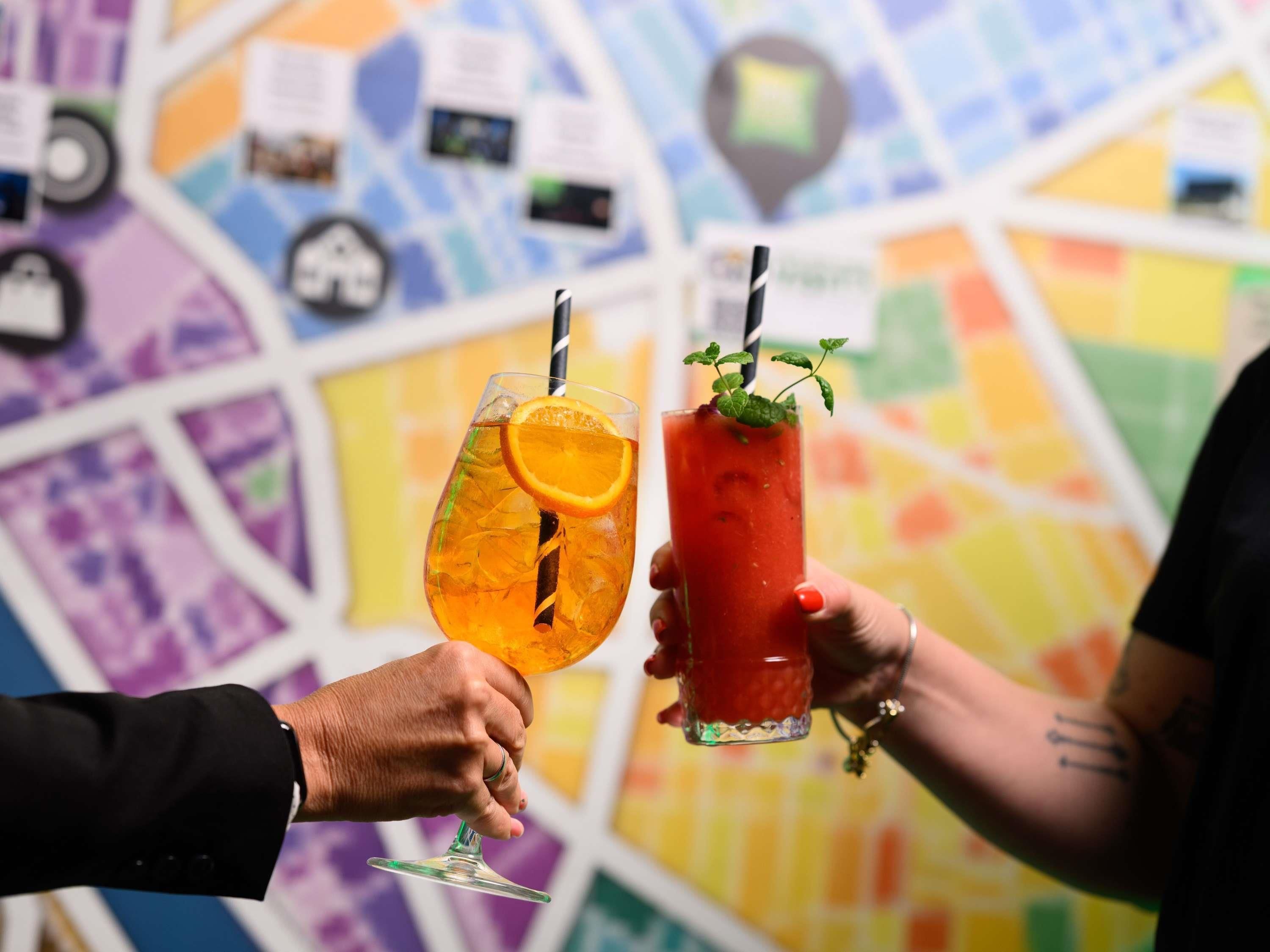 Two people holding glasses of drinks, one with a citrus garnish and the other with a mint garnish, against a colorful wall with various icons.