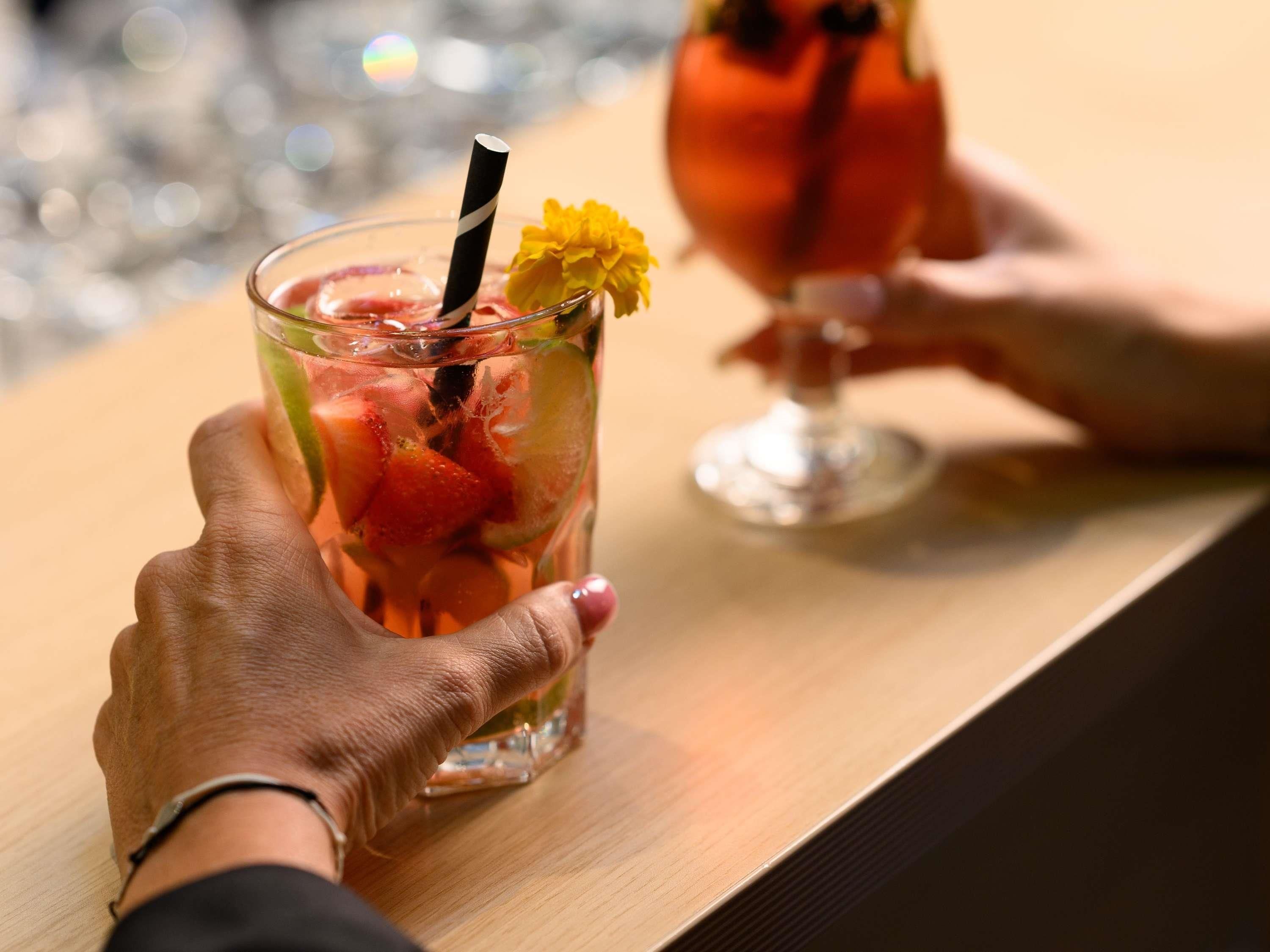 A person holding a glass with a straw and a flower, and another glass with a drink in the background.