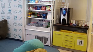 Habitación con kitchenette, refrigerador lleno de bebidas, microondas y cafetera. Hay dos puffs y una pared decorada con varios iconos.
