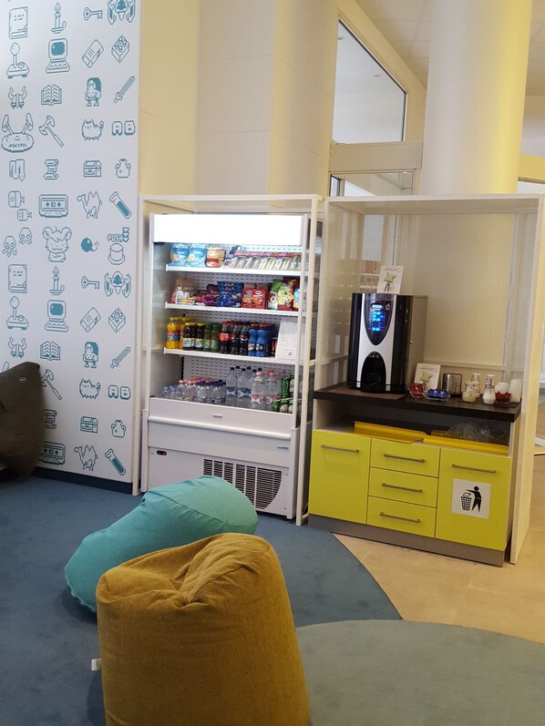 Habitación con kitchenette, refrigerador lleno de bebidas, microondas y cafetera. Hay dos puffs y una pared decorada con varios iconos.