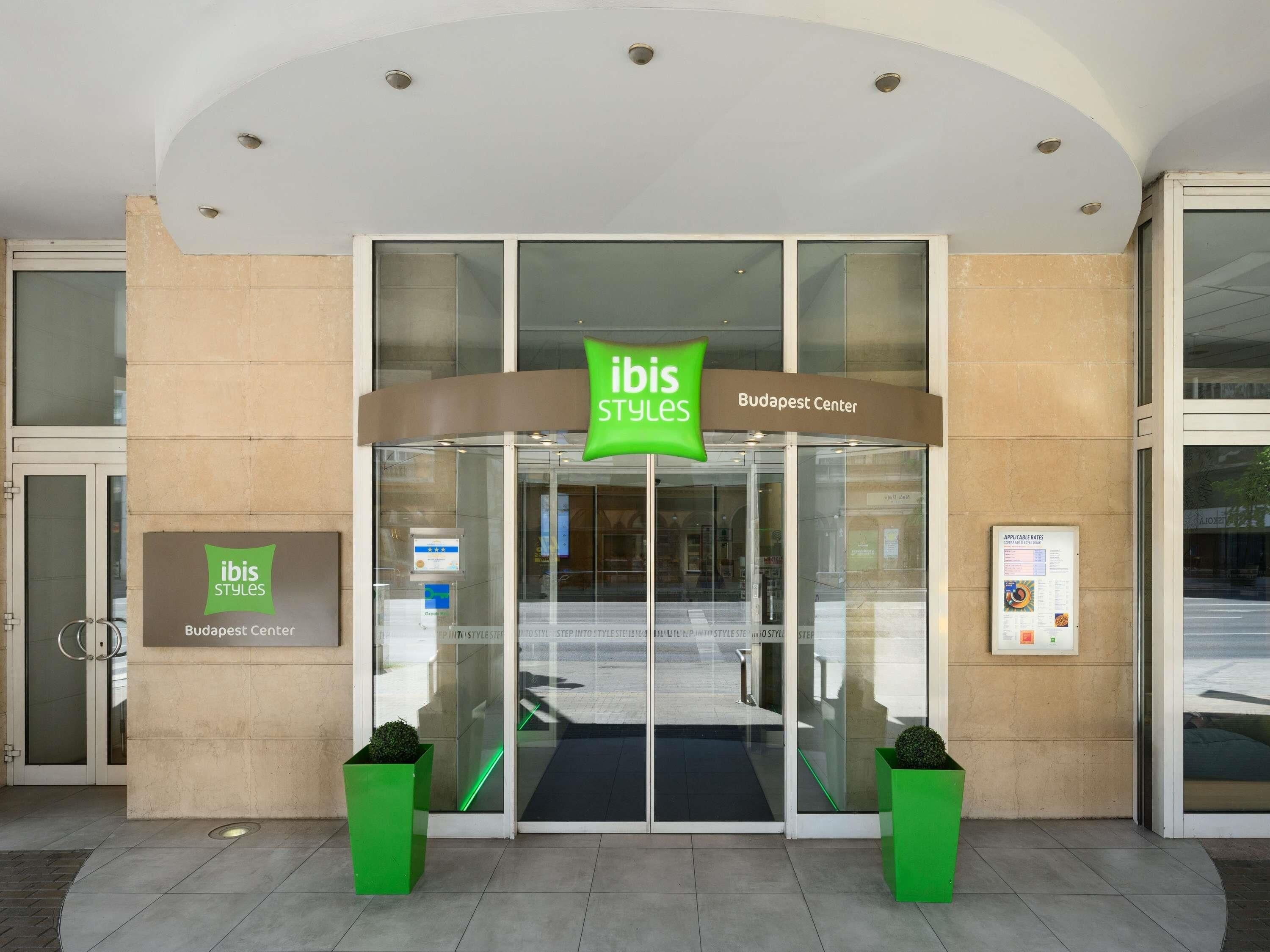 The entrance to ibis Styles Budapest Center with a glass door, signage, and two green planters.