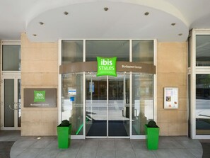 La entrada a ibis Styles Budapest Center con puerta de vidrio, señalización y dos macetas verdes.