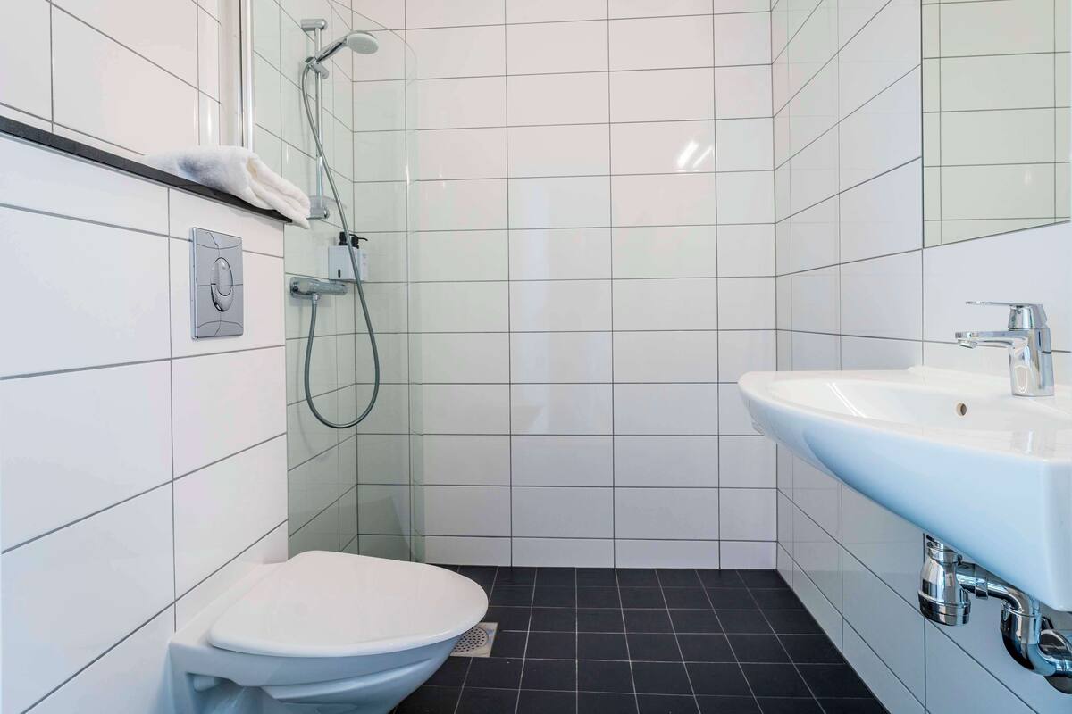 Une petite salle de bain moderne avec des murs recouverts de carreaux blancs, un sol noir, des toilettes blanches, une douche avec un pommeau réglable et un lavabo avec un robinet chromé.