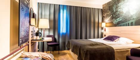 Habitación de hotel con cama, escritorio, silla, televisor y ventana grande con cortinas.