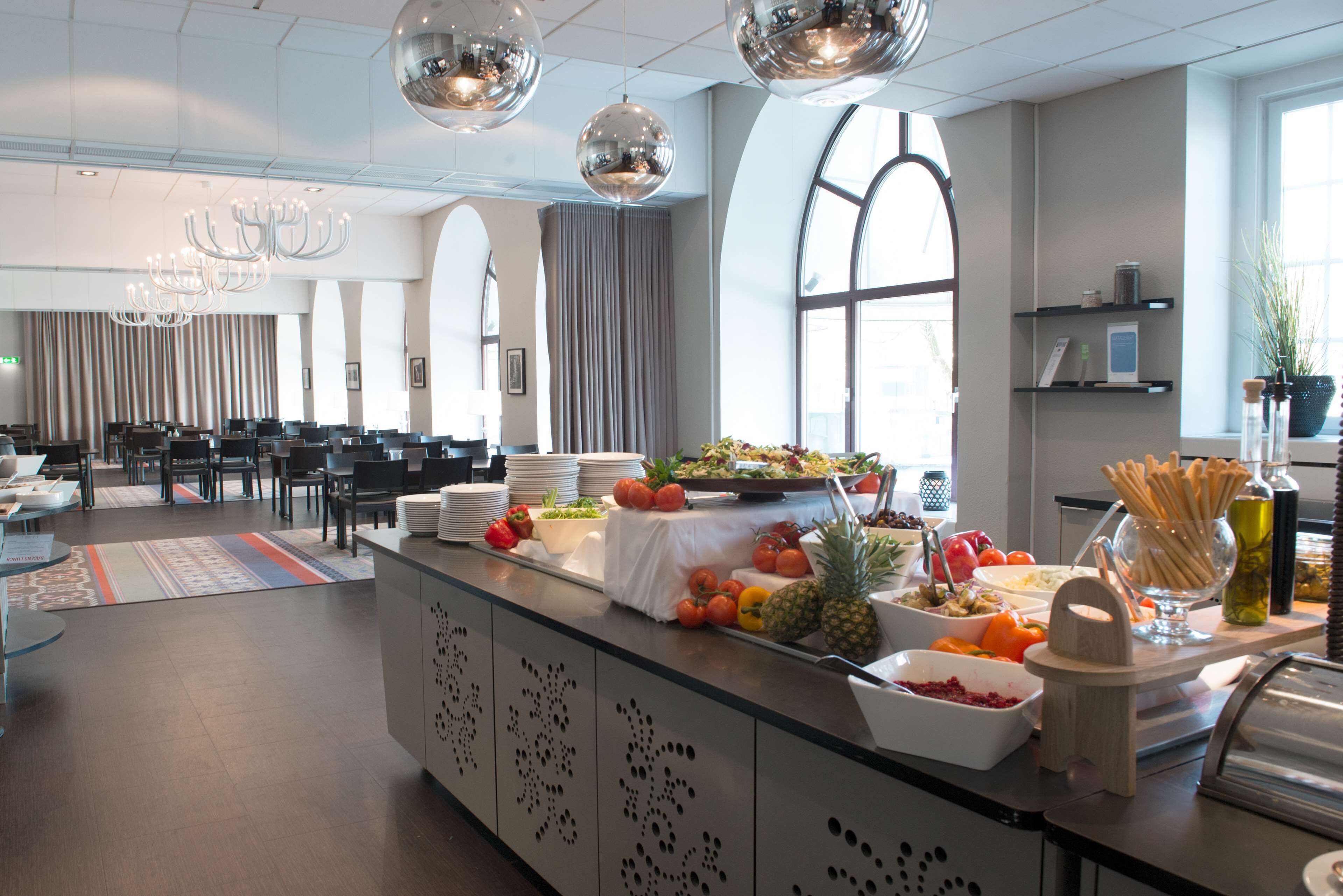 A modern hotel lobby with a buffet area featuring various fruits and vegetables, a seating area with tables and chairs, and a large window providing natural light.