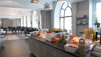 A modern hotel lobby with a buffet area featuring various fruits and vegetables, a seating area with tables and chairs, and a large window providing natural light.