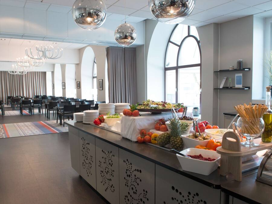 A modern hotel lobby with a buffet area featuring various fruits and vegetables, a seating area with tables and chairs, and a large window providing natural light.