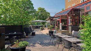 Un espacio al aire libre con mesas y sillas, rodeado de follaje y un edificio con ventanas grandes.
