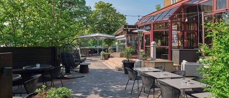 An outdoor patio area with tables and chairs, surrounded by greenery and a building with large windows.