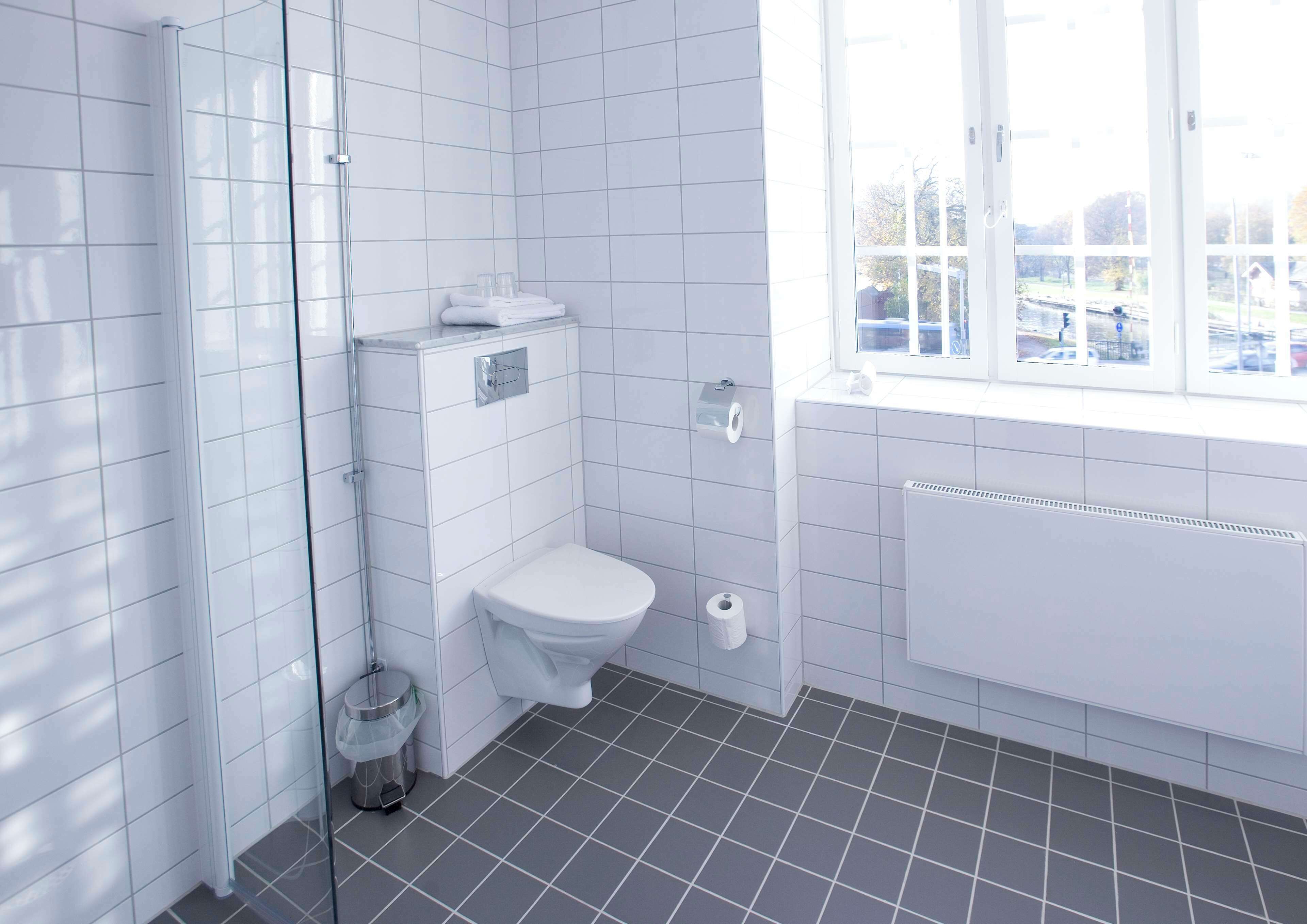 A modern bathroom with white tiles, a toilet, a glass shower enclosure, and a window with a view of the outside.