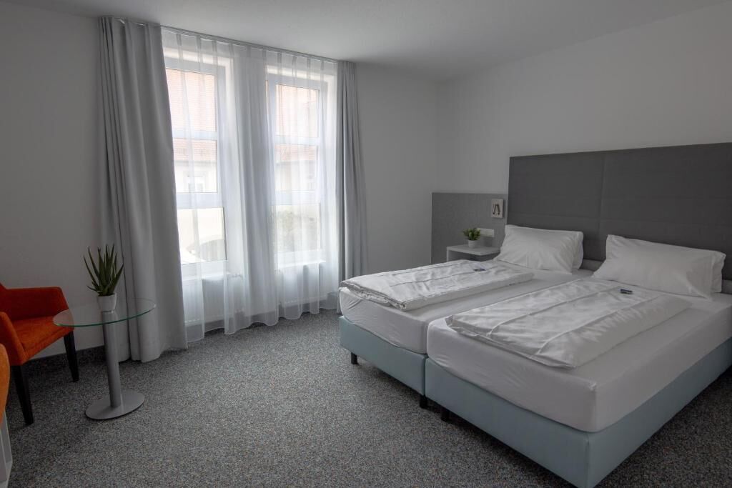 A hotel room with a bed, a chair, a small table with a plant, and a window with curtains.