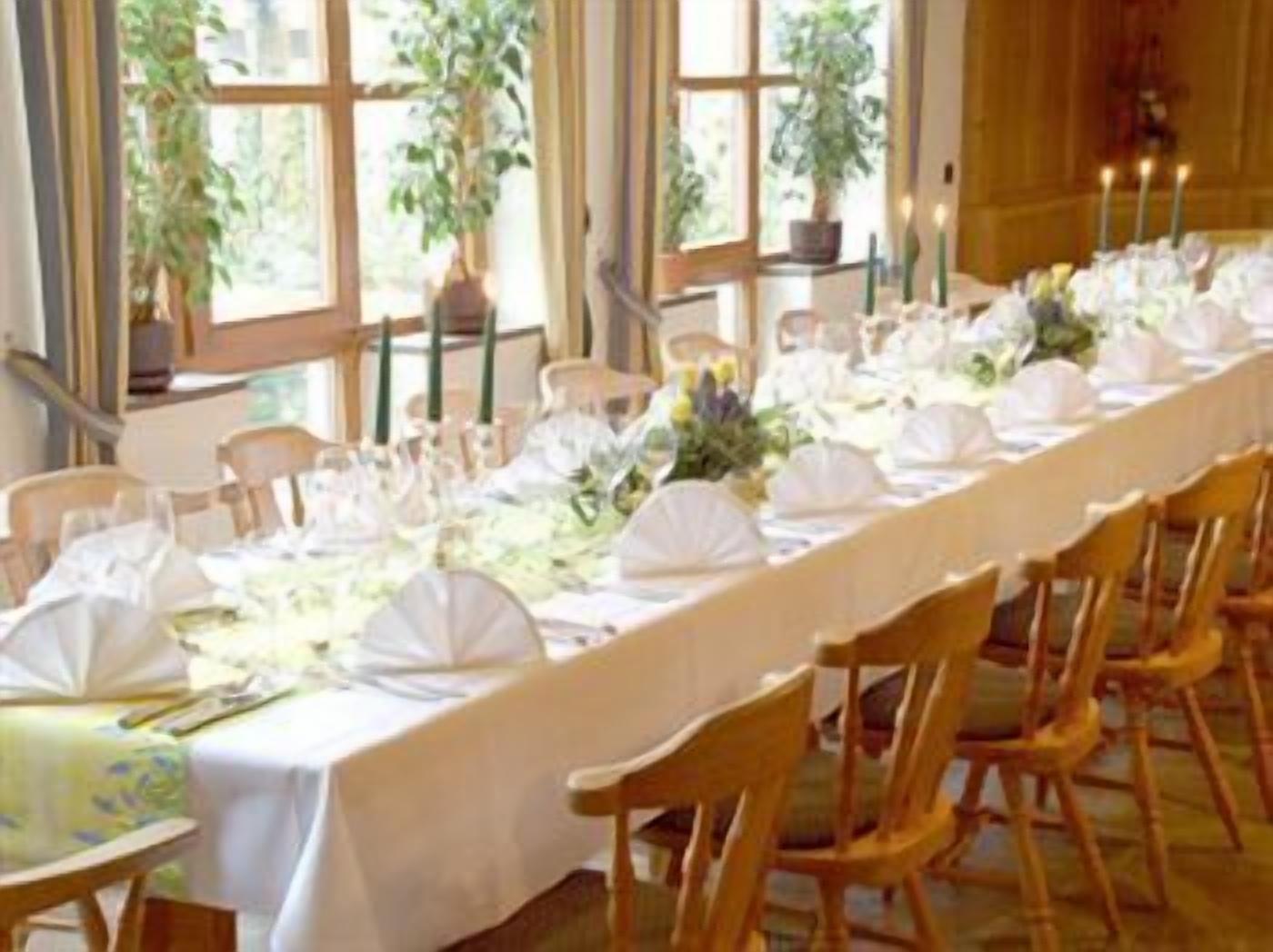 A long dining table set with white tablecloths, chairs, and candles, with large windows and potted plants in the background.