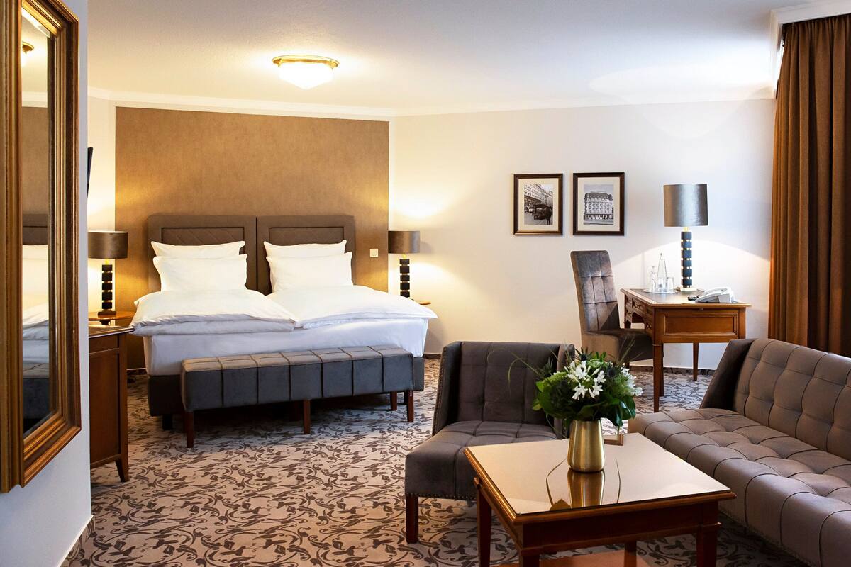 A hotel room with a large bed, a sofa, a small table, and a desk. There are two lamps, a mirror, and a framed picture on the wall.