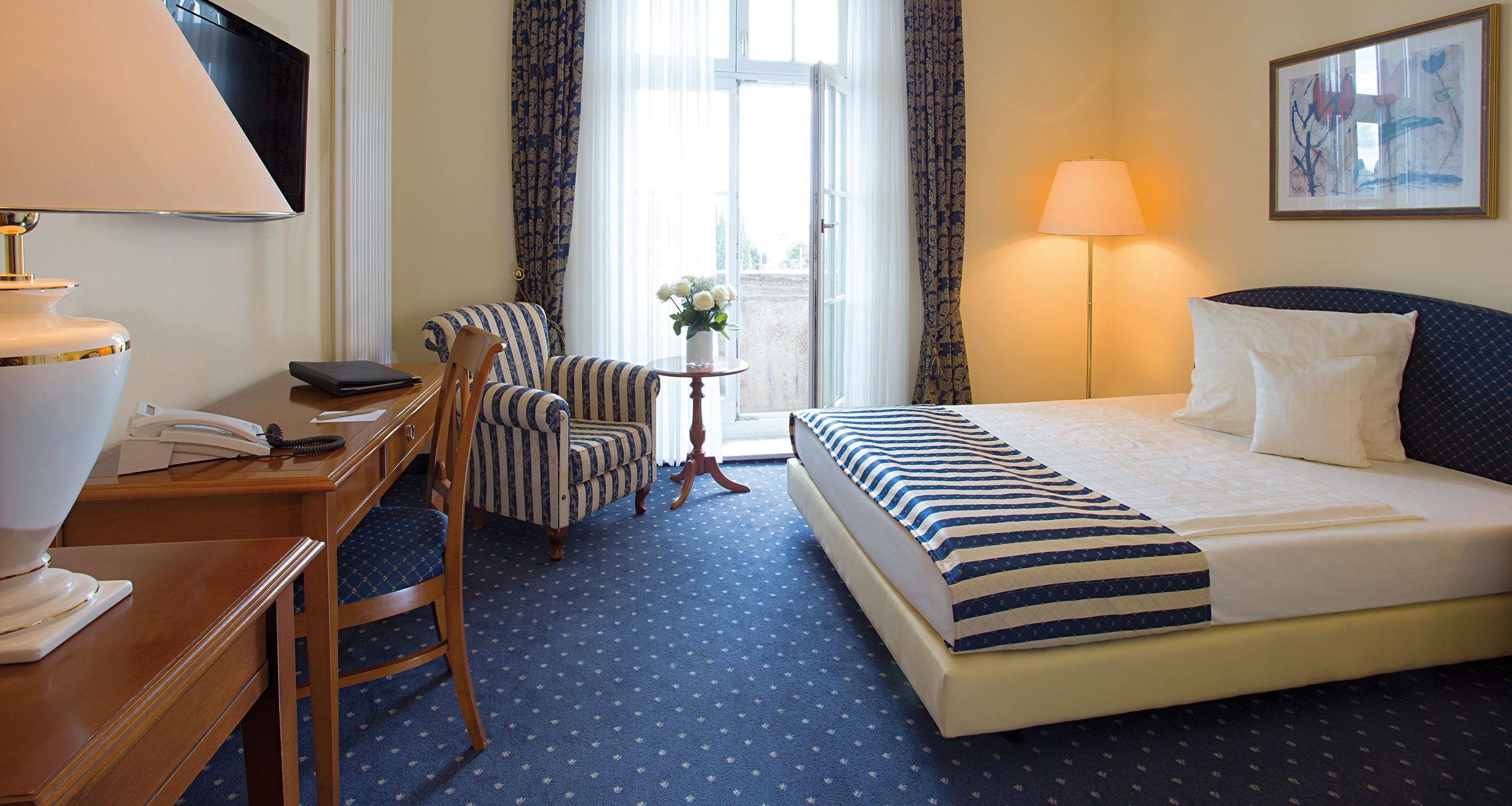 A hotel room with a bed, a desk with a telephone, a chair, a small table with a vase of flowers, and a lamp.