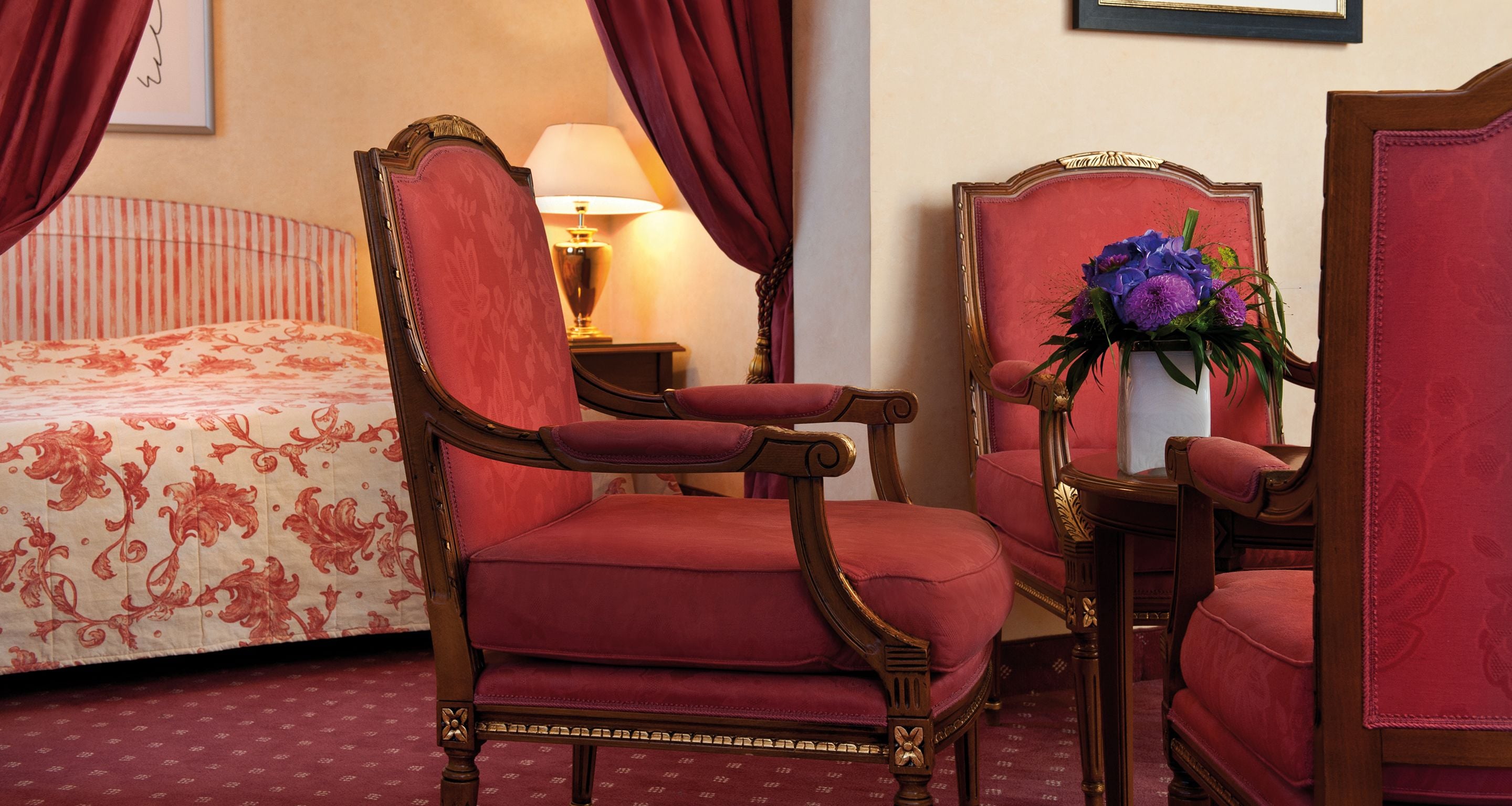 Een hotelkamer met een bed, een rode fauteuil, een houten nachtkastje en een vaas met bloemen.