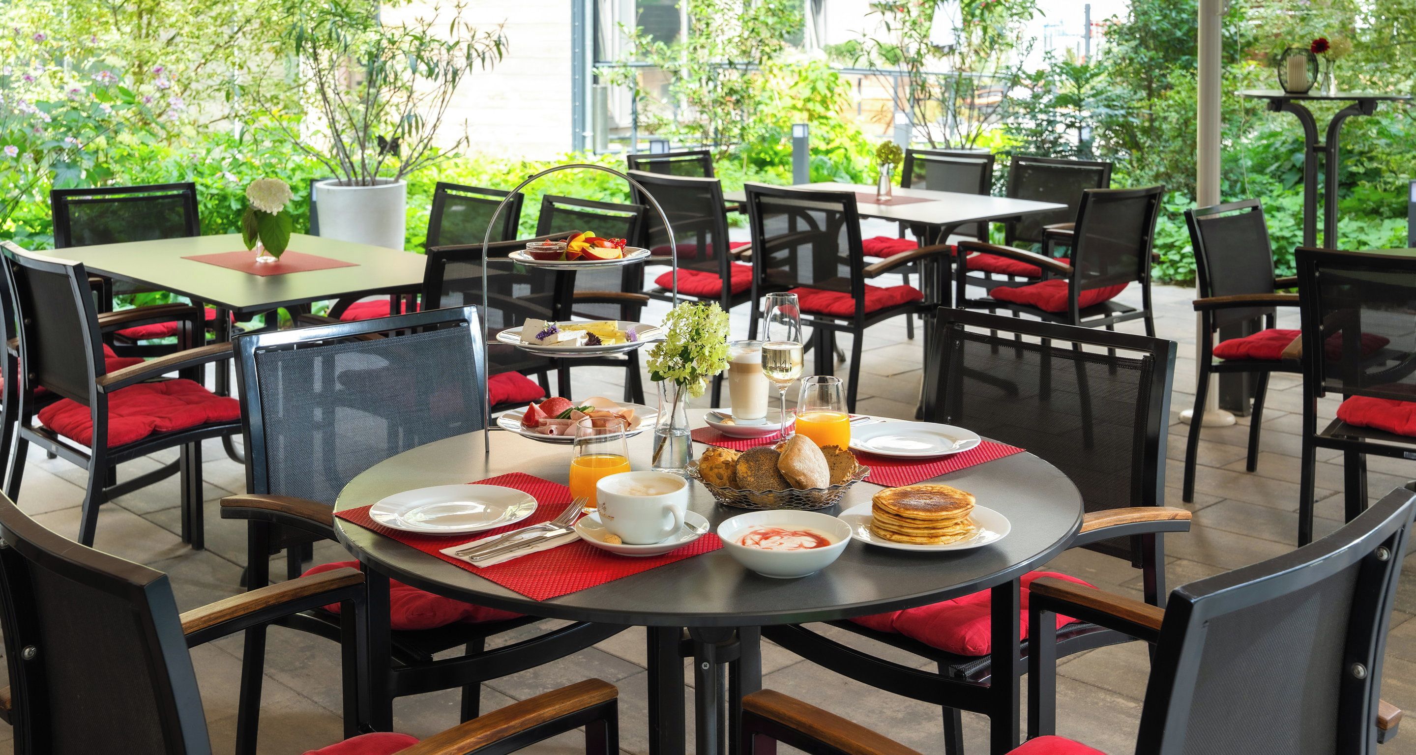 Une table de salle à manger ronde, entourée de chaises à coussins rouges, le tout agrémenté de verdure.