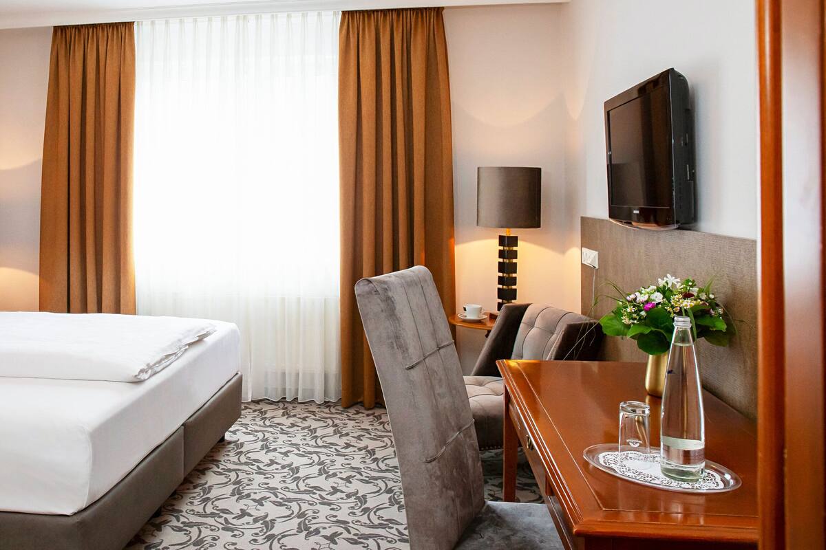 A hotel room with a bed, a desk, a chair, a TV, and a window with curtains.