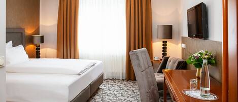 A hotel room with a bed, a desk, a chair, a TV, and a window with curtains.