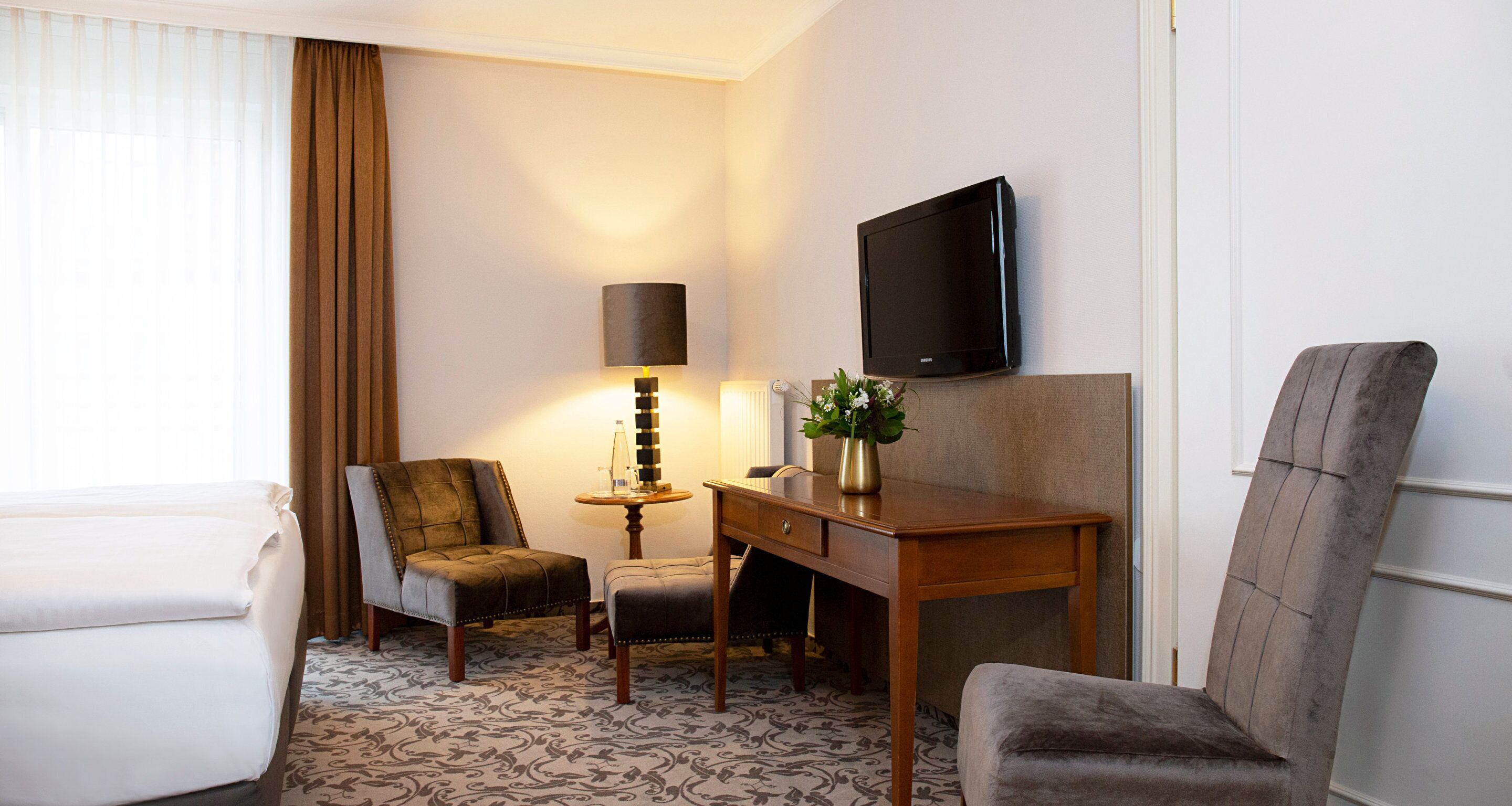 Ein Hotelzimmer mit einem Bett, zwei Sesseln, einem Schreibtisch, einer Lampe, einem Fernseher und einer Vase mit Blumen.