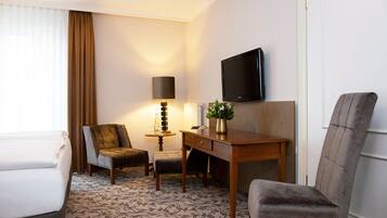 Ein Hotelzimmer mit einem Bett, zwei Sesseln, einem Schreibtisch, einer Lampe, einem Fernseher und einer Vase mit Blumen.