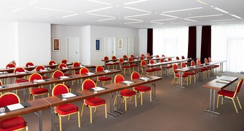 Un salón de conferencias con filas de mesas y sillas rojas, paredes blancas y ventanas grandes con cortinas.
