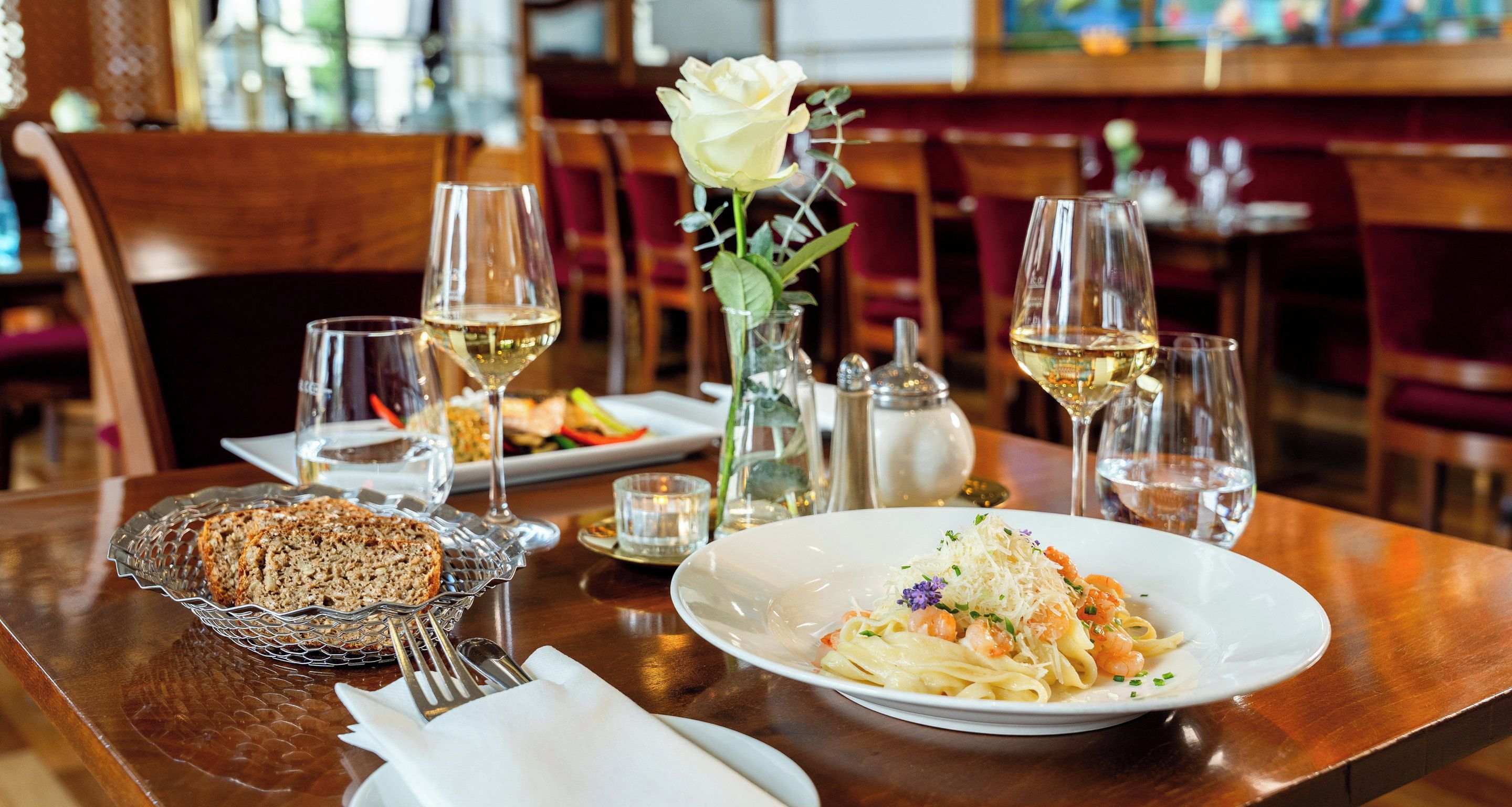 A dining table with a plate of pasta, a glass of white wine, and a bread basket.