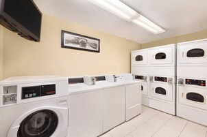 Une buanderie équipée de plusieurs machines à laver et de plusieurs sèche-linge, un téléviseur à écran plat fixé au mur et un tableau encadré au-dessus des machines.