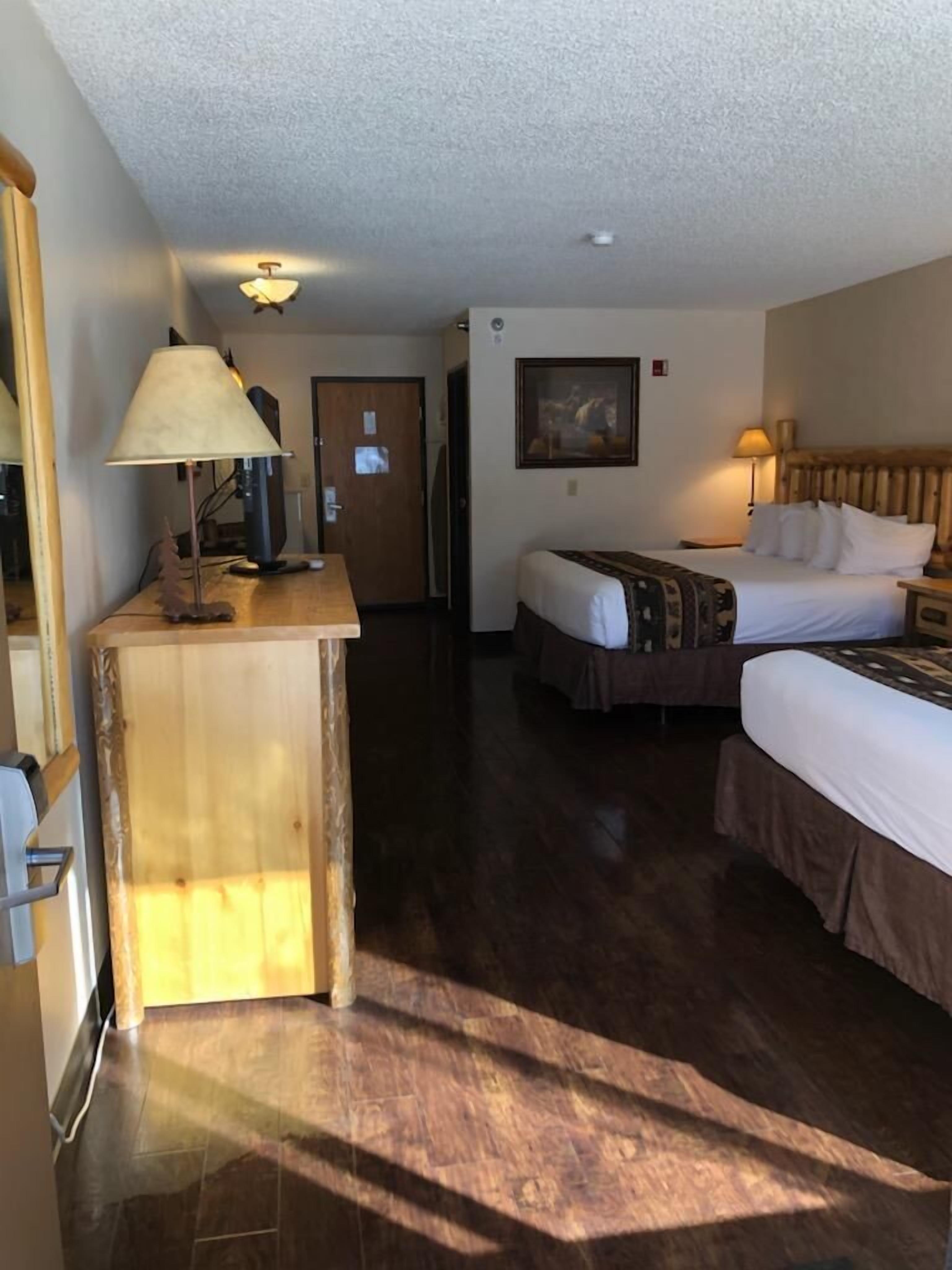 A hotel room with a wooden desk, a bed with a patterned bedspread, a nightstand with a lamp, and a painting on the wall.