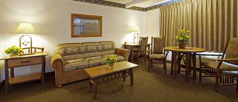 A cozy hotel room with a sofa, a coffee table, a dining table with chairs, lamps, and a framed picture on the wall.