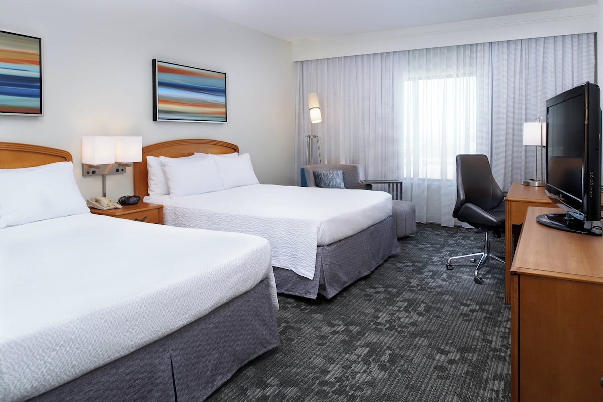 A hotel room with two beds, a desk with a TV, a chair, a lamp, and a window with curtains.