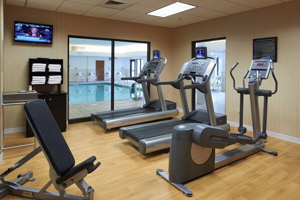 A gym with treadmills, an elliptical, and a bench. There is a television on the wall and a pool visible through the glass doors.