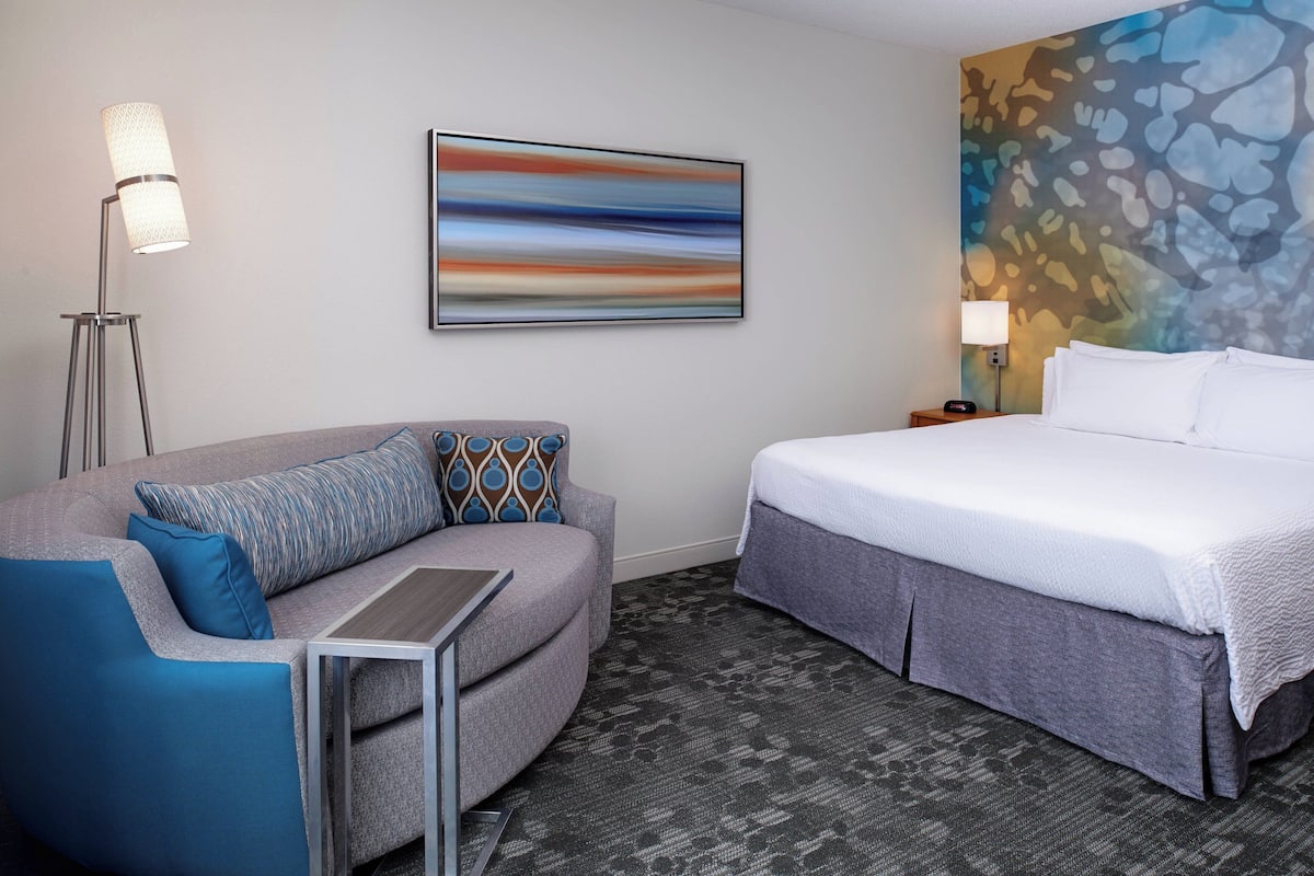 A hotel room with a bed, a sofa, a standing lamp, and a wall painting.